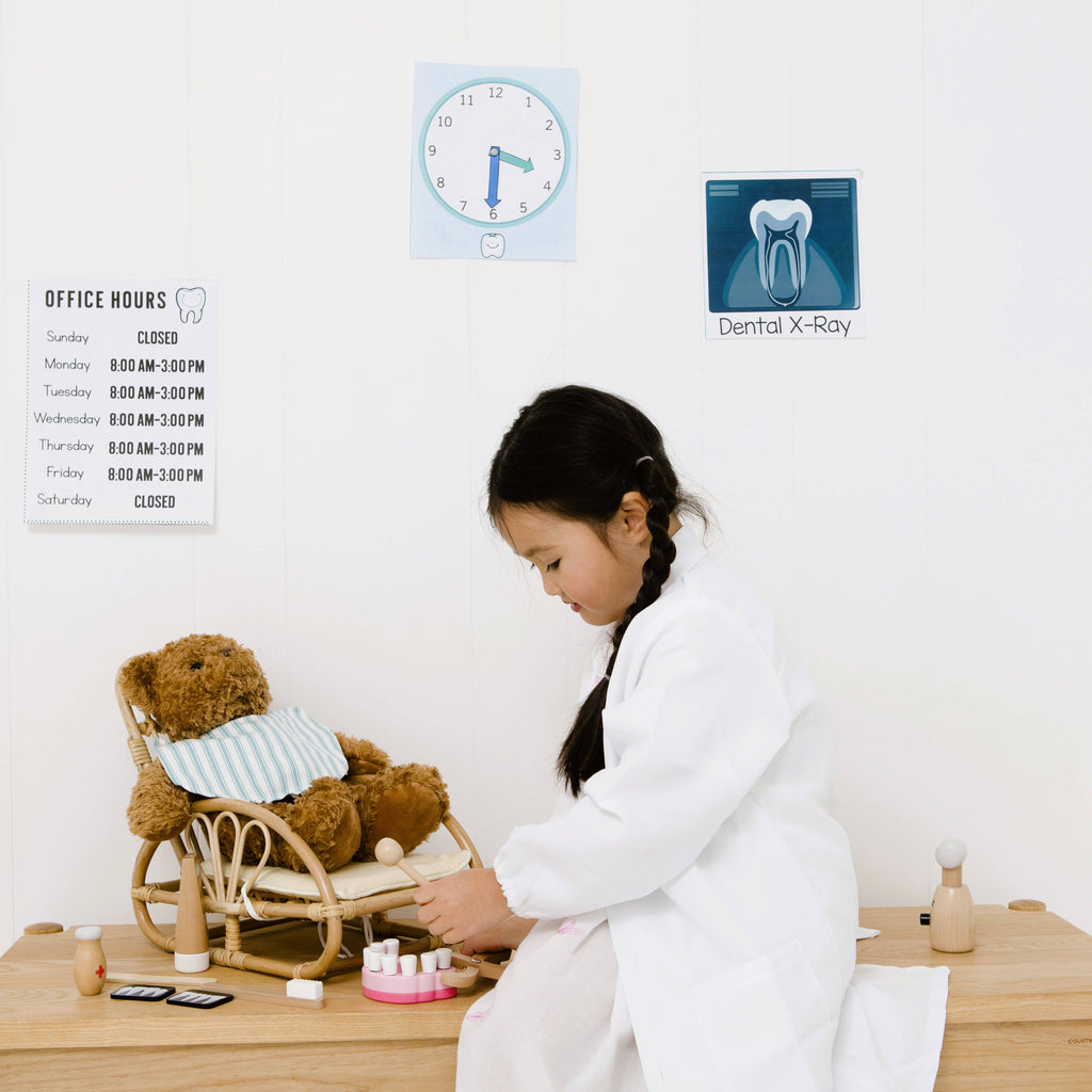 A young girl in a white coat pretends to be a dentist, examining a teddy bear with the Wooden Dentist Set. Dental posters, office hours, and toy dental tools from the set are arranged on the wall and table.