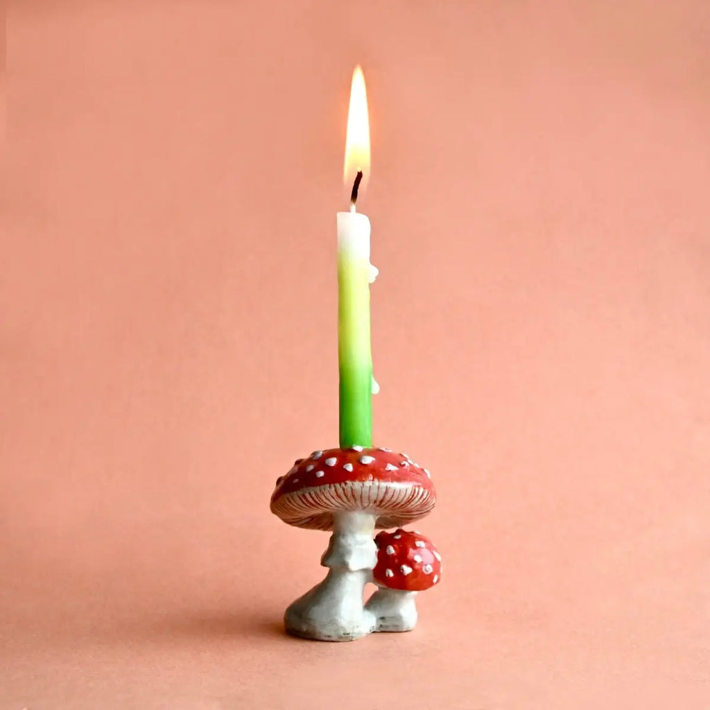 A lit green candle mounted on a hand-painted ceramic holder shaped like red and white spotted mushrooms, against a soft peach background.