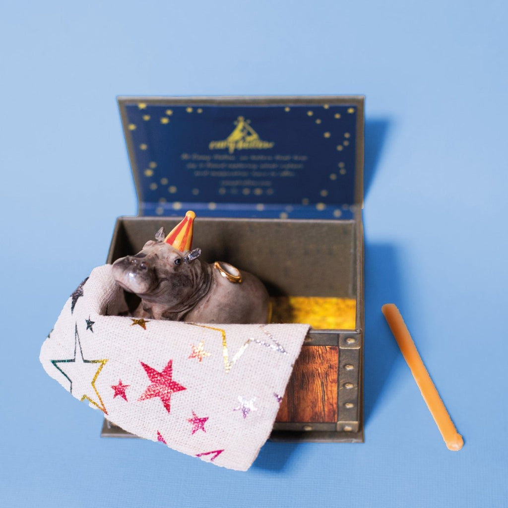 Sentence with product name: A small Hippo Cake Topper wearing a hand-painted party hat, nestled in an decorative open box with star-patterned fabric, against a blue background. A pencil lies next to the box.