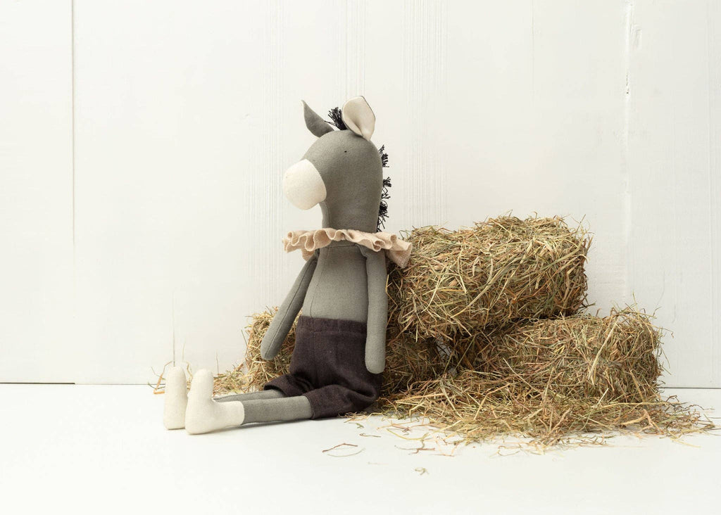 Introducing Darwin the Donkey, a plush toy made from recycled polyester, with gray fur, a tan ruffled collar, dark shorts, and small white feet. This cuddly companion lounges on hay bales in a simple white room.