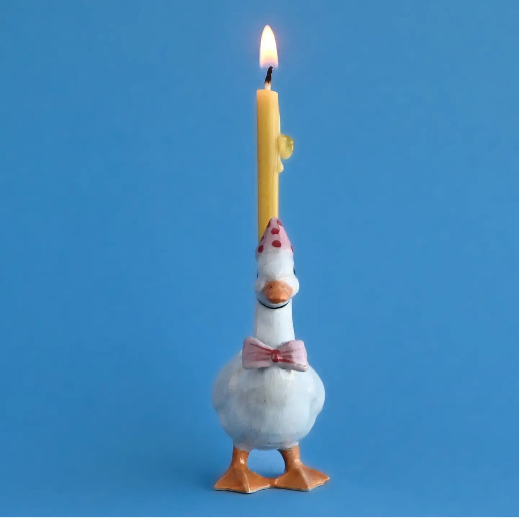 A whimsical Pink Goose Cake Topper wearing a pink bow tie and a polka dot party hat, with a lit yellow candle affixed to its back. The background is solid light blue.