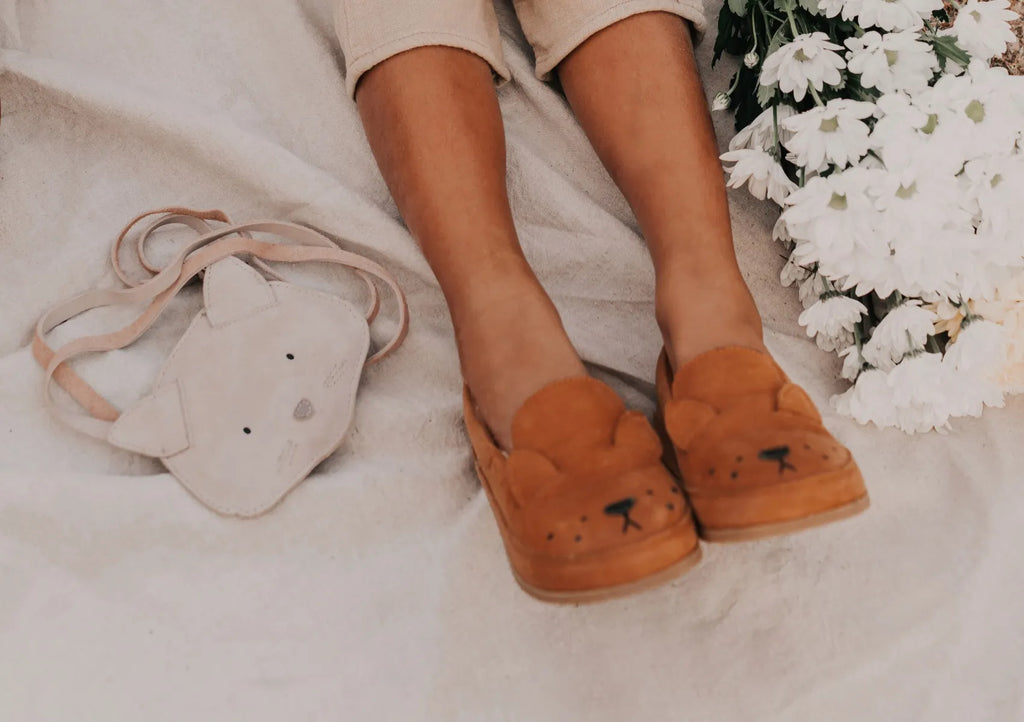 A close-up of a person's feet wearing cute bear-faced slippers, next to a Donsje Britta Classic Purse - Cat shaped like a bear's head, with white flowers in the background.