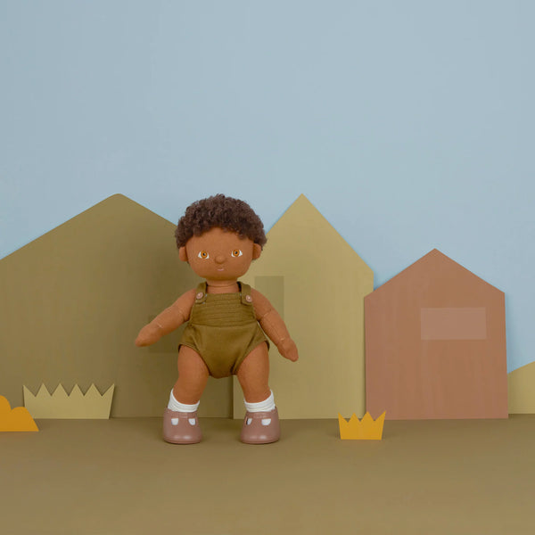 A Olli Ella Dinkum Doll with curly black hair and overalls sits between cut-out paper mountains and houses on a beige surface against a pale blue background.