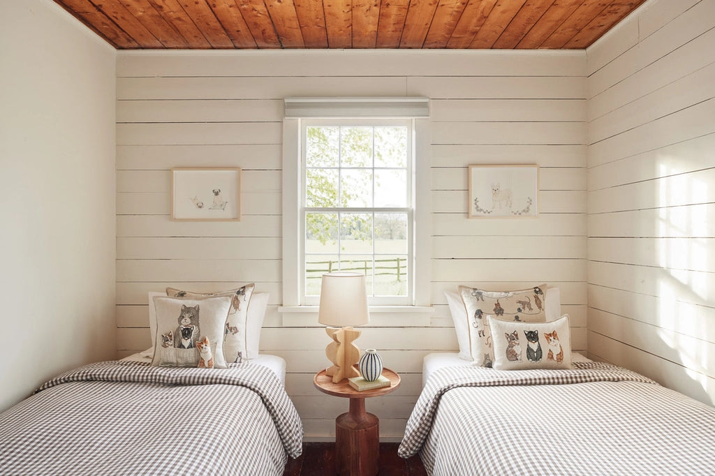 A cozy bedroom with a wood-paneled ceiling and walls, two twin beds with gingham bedding, Coral & Tusk Basket of Kittens Pocket Pillows, a central nightstand with a lamp, and framed animal prints on the walls.