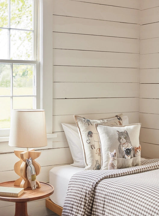 A cozy corner of a room featuring a bed with checkered bedding, a side table with a lamp and a wooden figurine, and decorative pillows including a Coral & Tusk Basket of Kittens Pocket Pillow, all against a backdrop of white