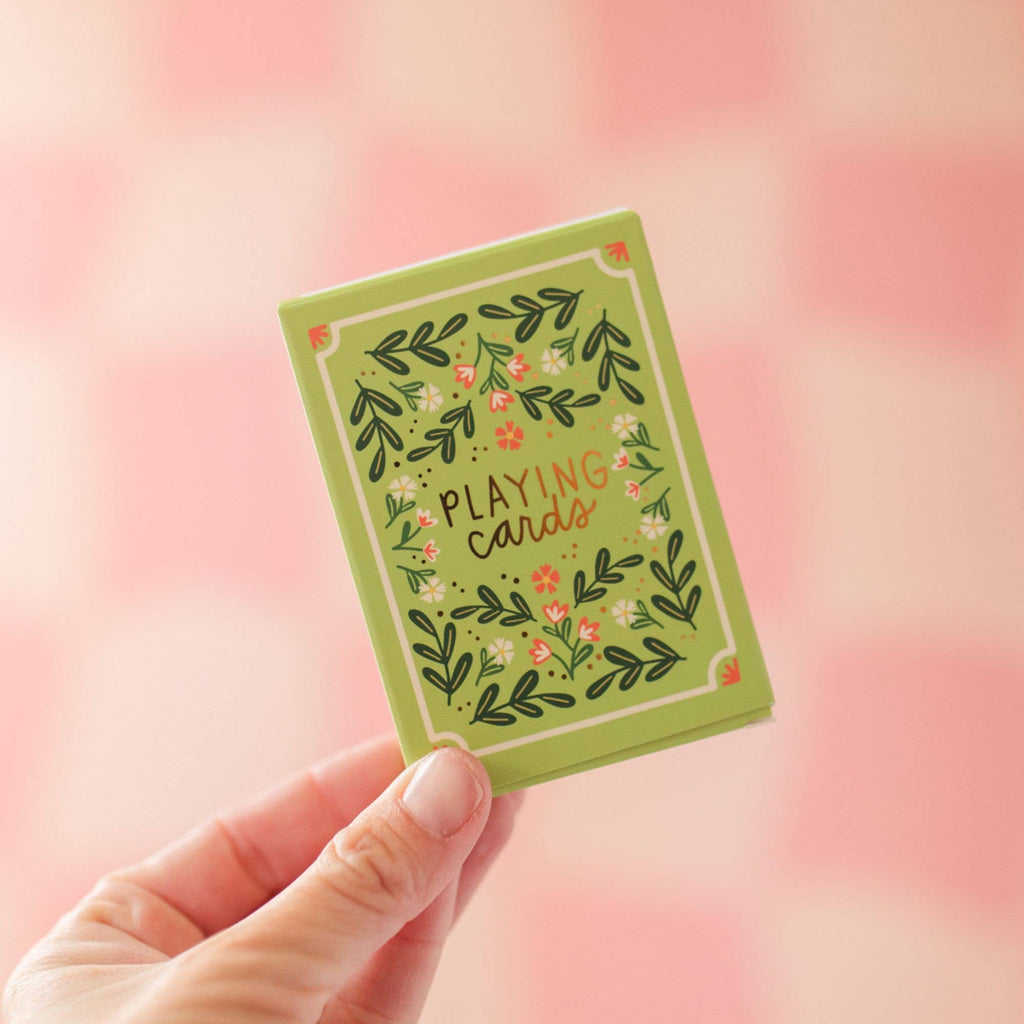 A hand holds the Green Floral Deck of Playing Cards (ships in approximately one week), featuring a hand-illustrated design and "Playing Cards" in the center, set against a soft pink checkered background—ideal for game lovers.