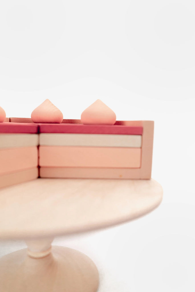 Three stacked pink books with peach-colored heart-shaped objects painted with non-toxic paint on top, displayed on a light wooden stand against a soft white background with faint circles.