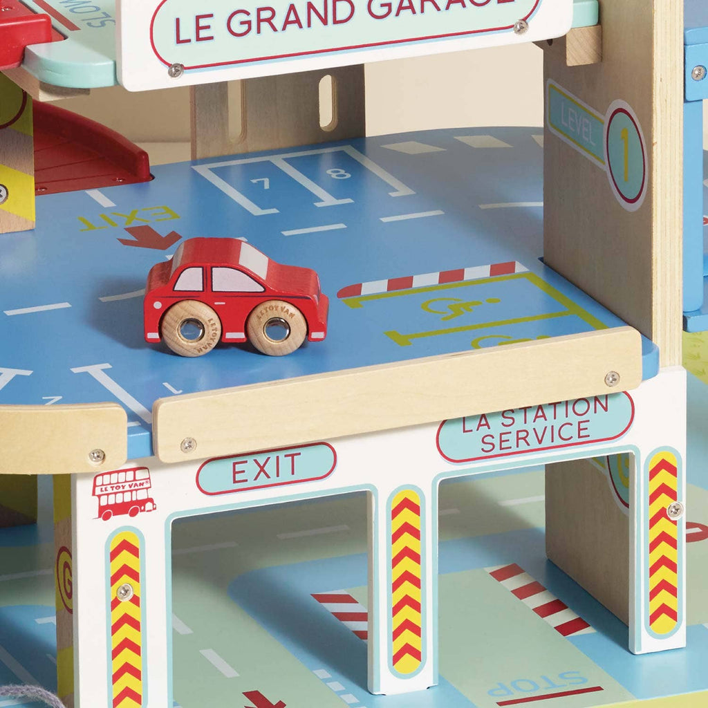 The Le Grand Wooden Garage inspires imaginative play, featuring a red car on a blue level, parking spaces, a ramp, and signs reading “LE GRAND GARAGE,” “EXIT,” and “LA STATION SERVICE.”.