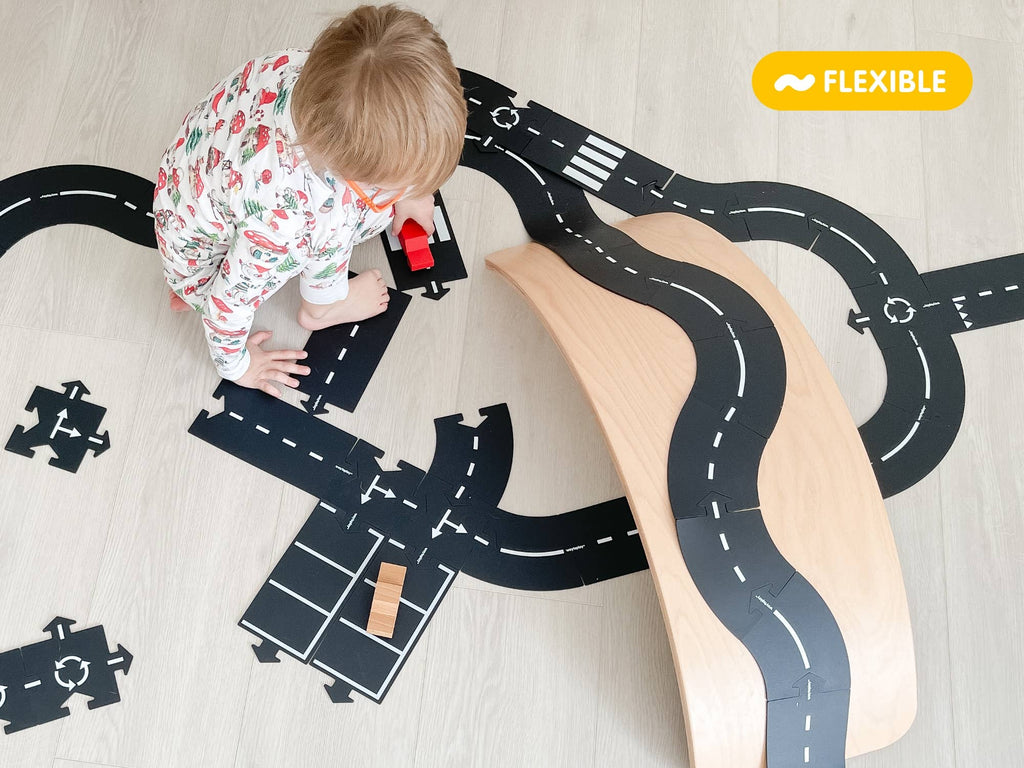 A young child in pajamas plays on the floor with the WaytoPlay: Extra Long Flexible Road Set - King of the Road, moving a red car along black road pieces and over a curved wooden ramp.