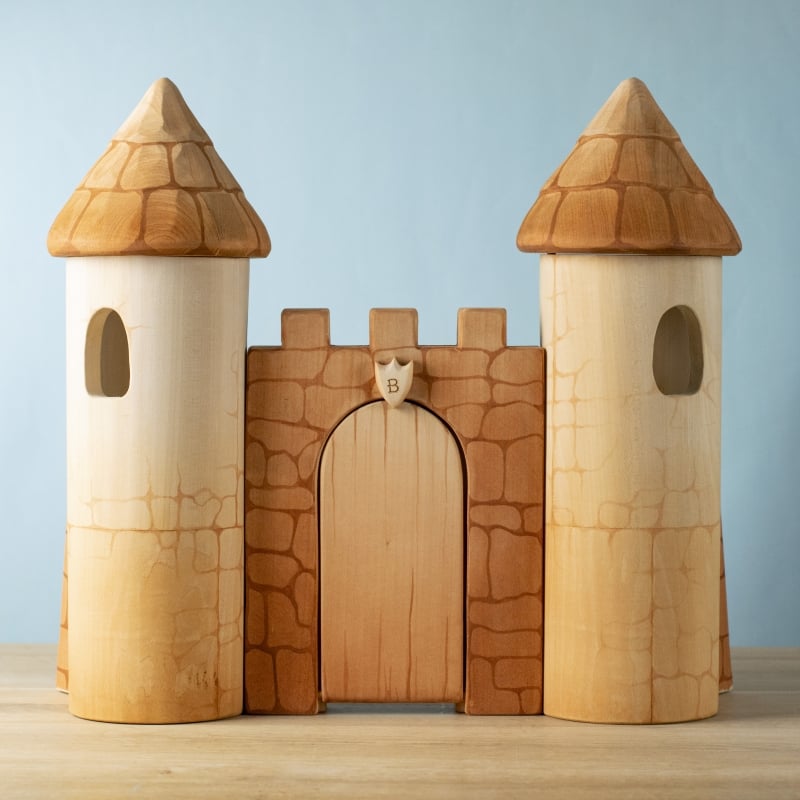 The Bumbu Handmade Wooden Castle is crafted from sustainable wood, with two tall cylindrical towers and a central arched gate featuring a carved stone pattern, set on a light wooden surface against a blue background.