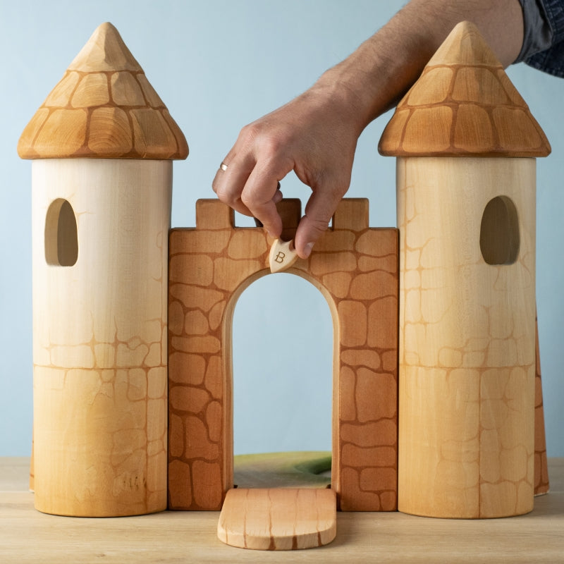 A hand places a "B" block atop the arched entrance of the Bumbu Handmade Wooden Castle, featuring two towers and a drawbridge, all made from sustainable wood and set on a wooden surface.