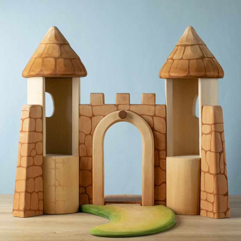 The Bumbu Handmade Wooden Castle features sustainable wood, two round towers, a central arched gate, stone-like patterns, and a green curved path at the entrance, all set against a light blue background.