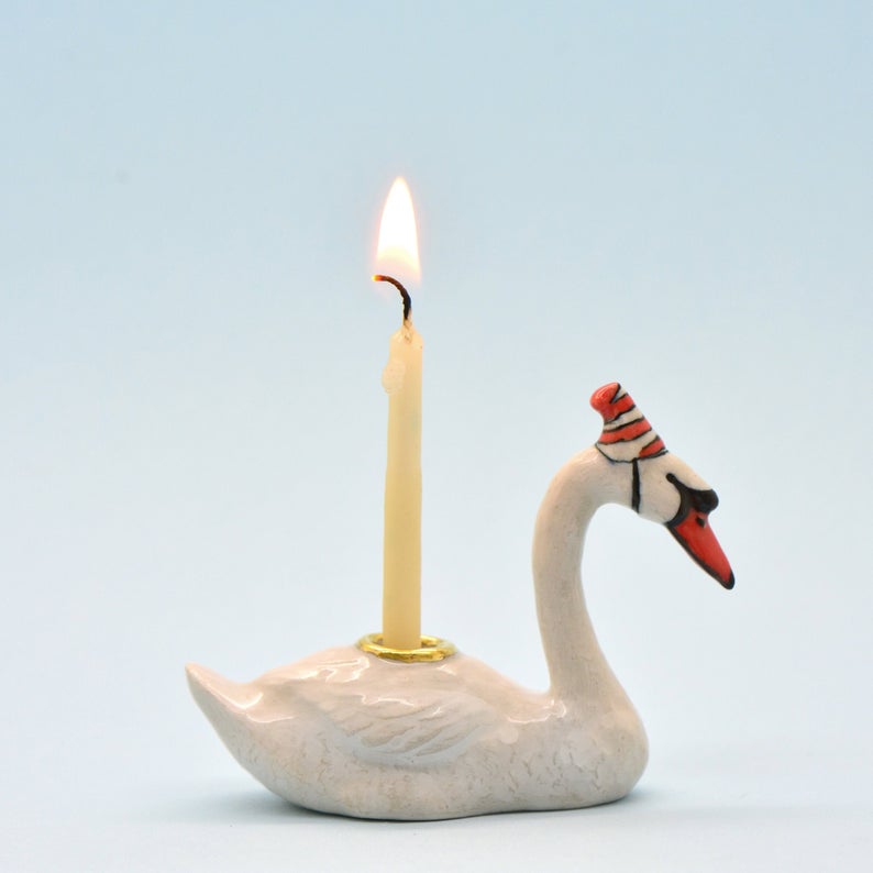 A hand-painted Swan Cake Topper, shaped like a swan and designed to hold a candle, is set against a plain light blue background. The swan features delicate details and a painted beak.