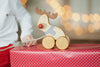 A child in a white shirt and red pants holds a string attached to a wooden playmate, the Handmade Wooden Reindeer Pull Toy. The toy has wheels, a painted blue scarf, and a red nose. It sits on a red polka-dotted gift box, with blurred holiday lights in the background.