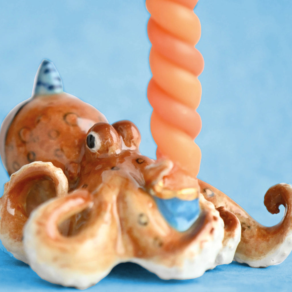 A fine porcelain Octopus Cake Topper with a textured body and swirling tentacles, holding an orange spiral shell against a soft blue background.