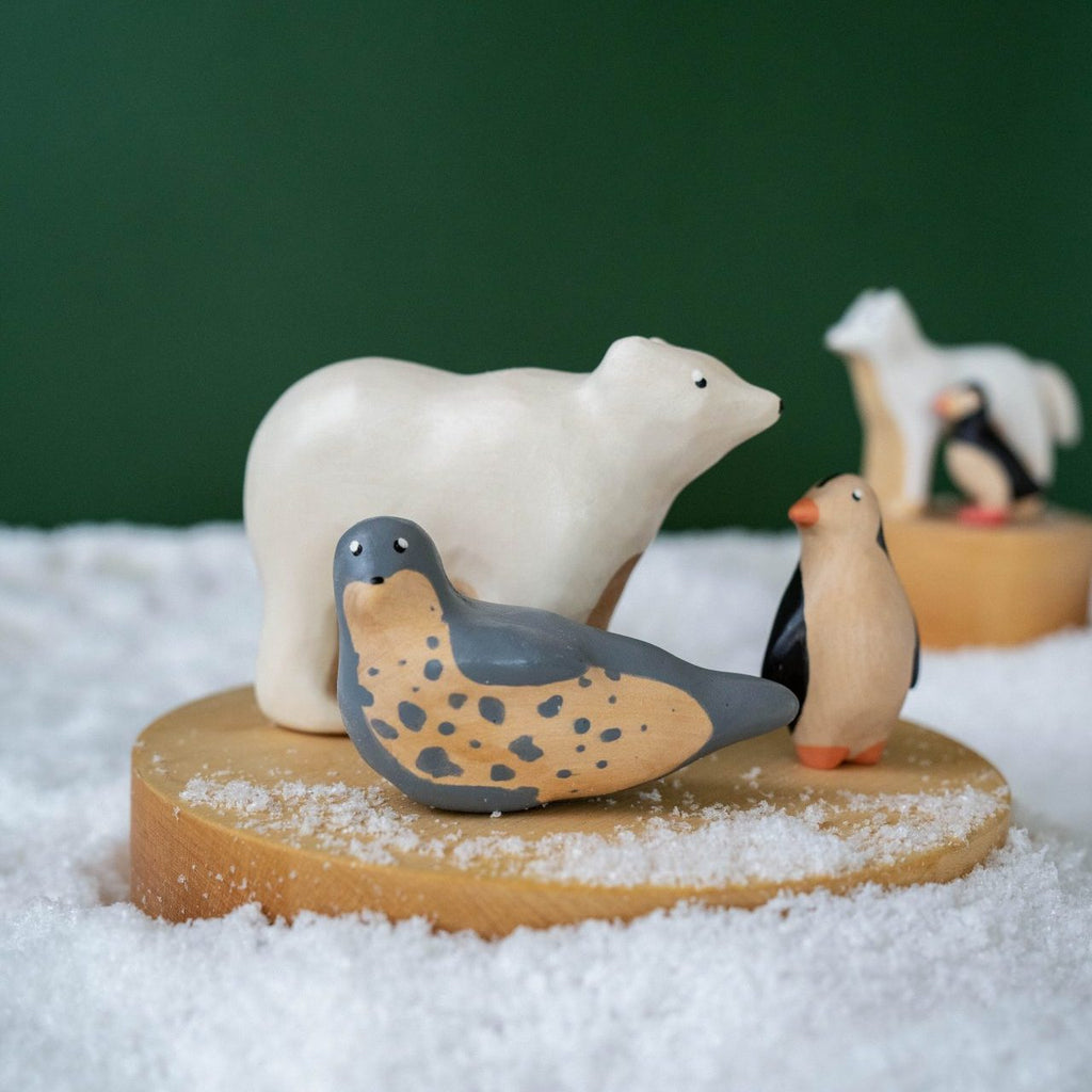 A collection of ceramic animal figurines, including a polar bear, a seal, and two penguins, displayed on a Handmade Wooden Arctic Wolf coated with non-toxic child-safe paint amidst artificial snow against a green