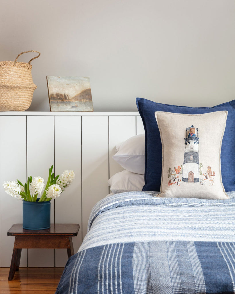 A cozy bedroom corner featuring a blue and white striped bedspread, a small wooden stool with a blue pot of white flowers, a Coral & Tusk Lighthouse Friends Pocket Pillow pillow with a lighthouse design, and a woven basket.