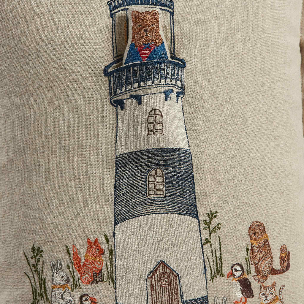 Detailed embroidery on a Coral & Tusk Lighthouse Friends Pocket Pillow depicting a lighthouse with a lighthouse keeper bear in the window, surrounded by various animals like foxes, rabbits, and birds in a serene scene.