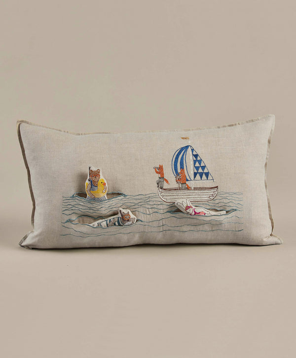 A Coral & Tusk Swimmers Pocket Pillow featuring an embroidered design of cats on a sailboat and floating on inner tubes, set against a neutral beige background, ideal for ocean-themed decor.