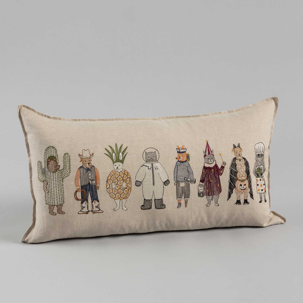 Sentence with Product Name: A Trick or Treat Pillow featuring illustrations of various whimsical characters in Halloween costumes, including a cactus, a cowboy, and a pineapple among others on a neutral-colored background.