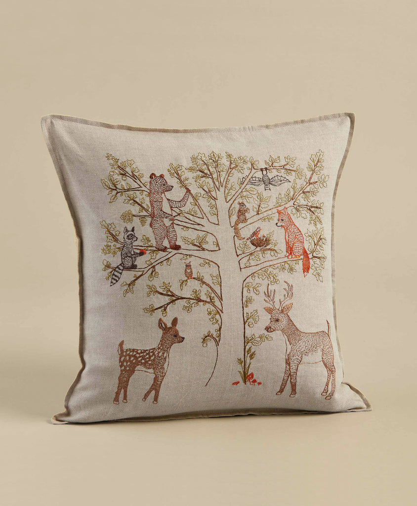 The Coral & Tusk Woodland Living Tree Pillow is a square beige cushion embroidered with a family tree and forest animals—bear, raccoon, fox, owl, and deer—arranged on the branches. Ships in one week.