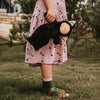 A young child in a pink dress adorned with cherry prints holds an Olli Ella Cozy Dinkum Doll - Black Cat Nox. The focus is on the doll and the child’s hands, with grass and a house faintly visible.
