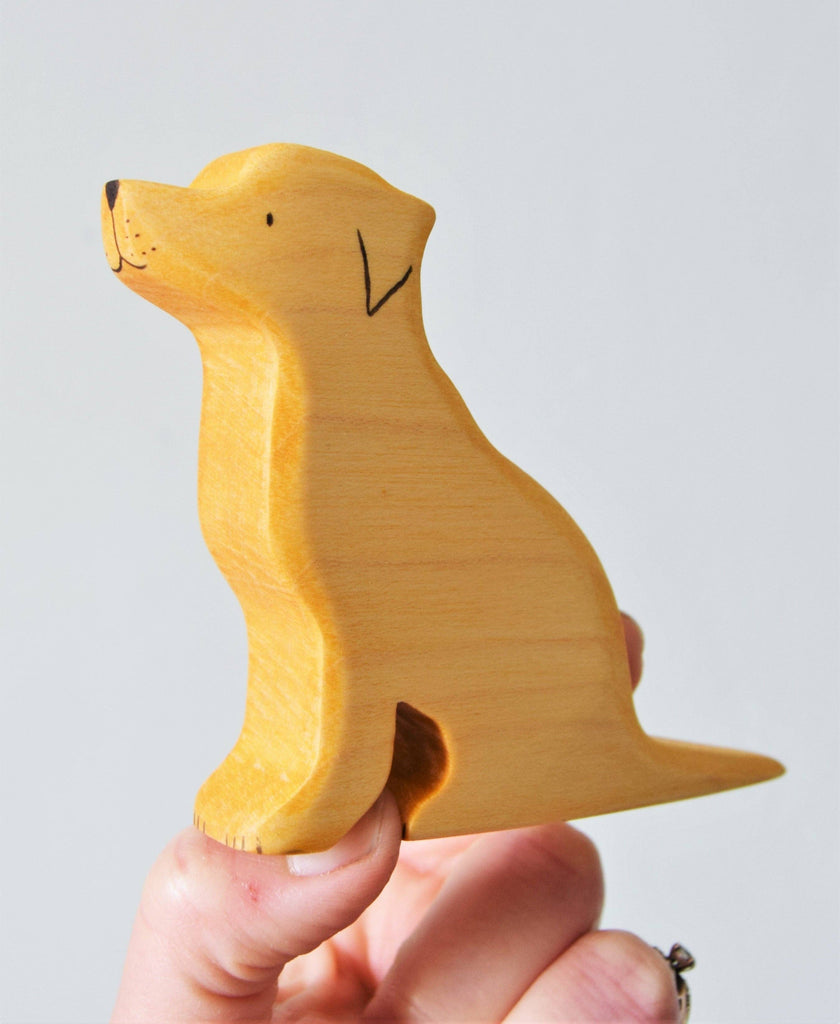 A hand holds the Handmade Wooden Labrador Dog, a small figurine carved from sustainable hardwood. It features a smooth surface with minimal facial details, enhanced by natural plant-based stains that highlight the wood's light, natural color.