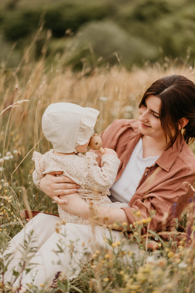 A woman smiles at a small child in a bonnet and floral outfit on her lap, surrounded by tall grass and wildflowers. The cozy scene is complete as the child gently holds a Sabo Concept Bear Rattle - Natural from Ukraine.