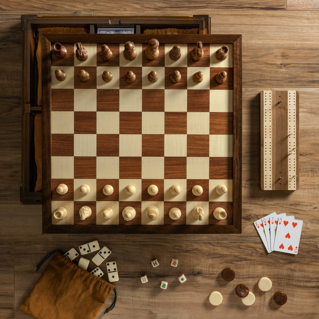 The Heirloom Edition: 7-in-1 chessboard is displayed with pieces ready for play, surrounded by dominoes, playing cards, a cribbage board, and a drawstring pouch with game pieces on a wooden surface.