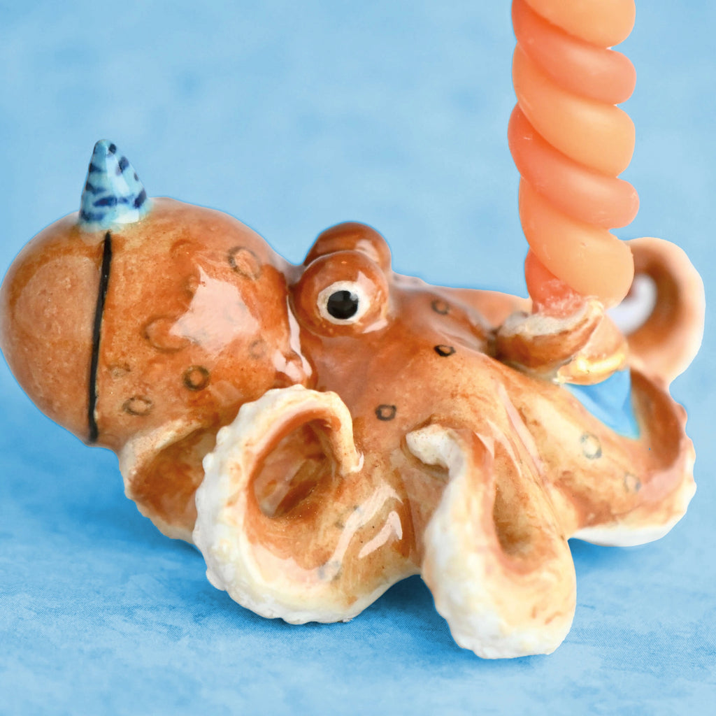 A Octopus Cake Topper with a textured, orange and cream body, holding a pink spiral object. The hand-painted octopus features detailed eyes and suction cups, set against a light blue background.