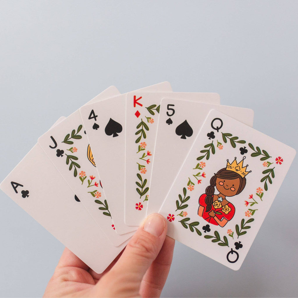 A hand displays five spade cards—ace, jack, four, king, and five—plus a queen of spades with a cartoon woman in a crown and floral border. The Green Floral Deck of Playing Cards ships in about one week. Ideal for fans of hand-illustrated design.