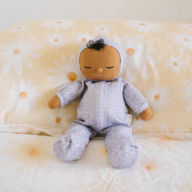 A Olli Ella Dozy Dinkums - Squeak from the limited-edition Daydream Collection with a brown complexion, wearing a lavender onesie with a dotted pattern, and a cute bunny-eared hood, sits on a pillow.