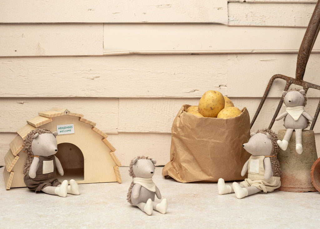 Harold The Hedgehog plush toys wear cloth outfits and pose by their charming wooden house. A paper bag of potatoes and a garden fork nearby hint at secret garden adventures against a light-colored wall, making them perfect companions for imaginative play.