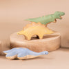 Three handmade wooden plesiosaurus dinosaur toys on a beige background. One resembling a t-rex stands upright, another shaped like a stegosaurus is on a wooden base, and a third resembling a br