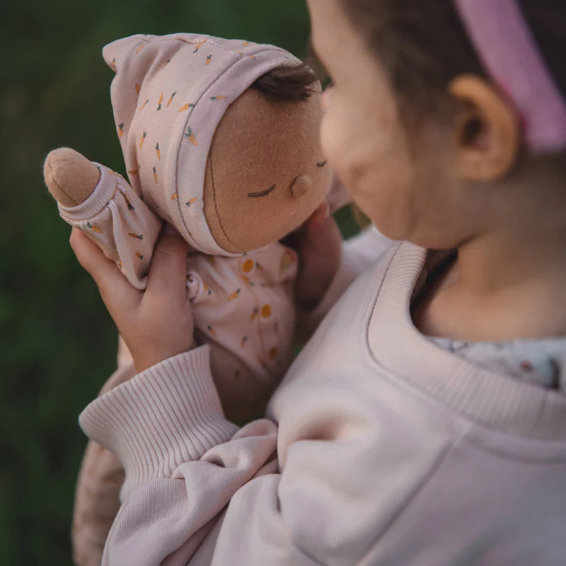 A young girl gently holding a Olli Ella x Odin Parker Dozy Dinkum - Bugsy Hopscotch cloth doll with a matching floral hat to her cheek, showcasing a tender and affectionate moment.