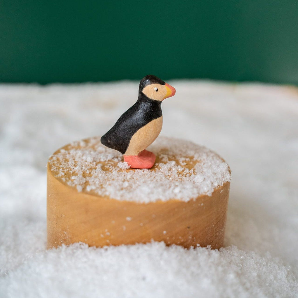 A small handcrafted wooden puffin figurine stands on a round wooden block surrounded by artificial snow, against a dark green background.