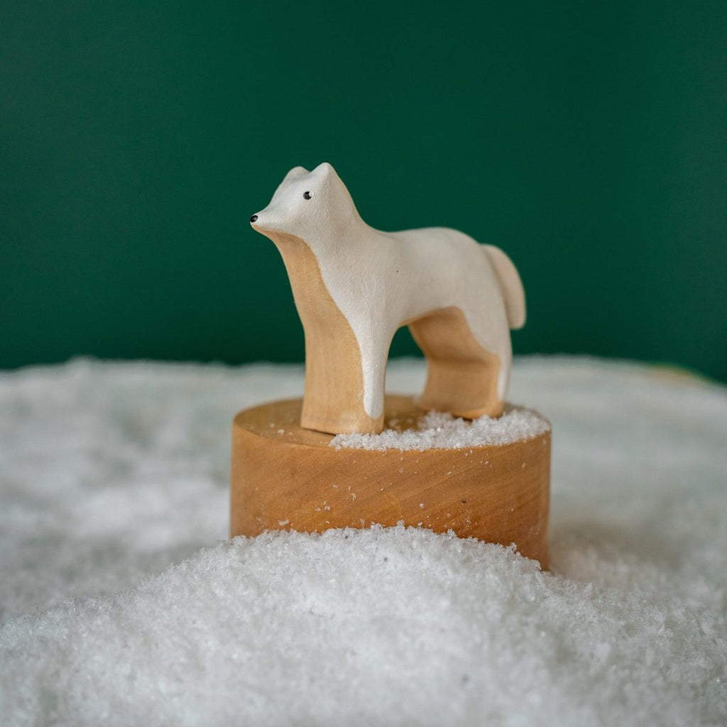 A handmade wooden Arctic Wolf figurine standing on a circular wooden base surrounded by artificial snow, handcrafted in Serbia, set against a deep green background.