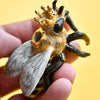 A hand holding a Queen Bee Cake Topper with a golden crown, black and white body, and intricately hand-painted wings, set against a bright yellow background.