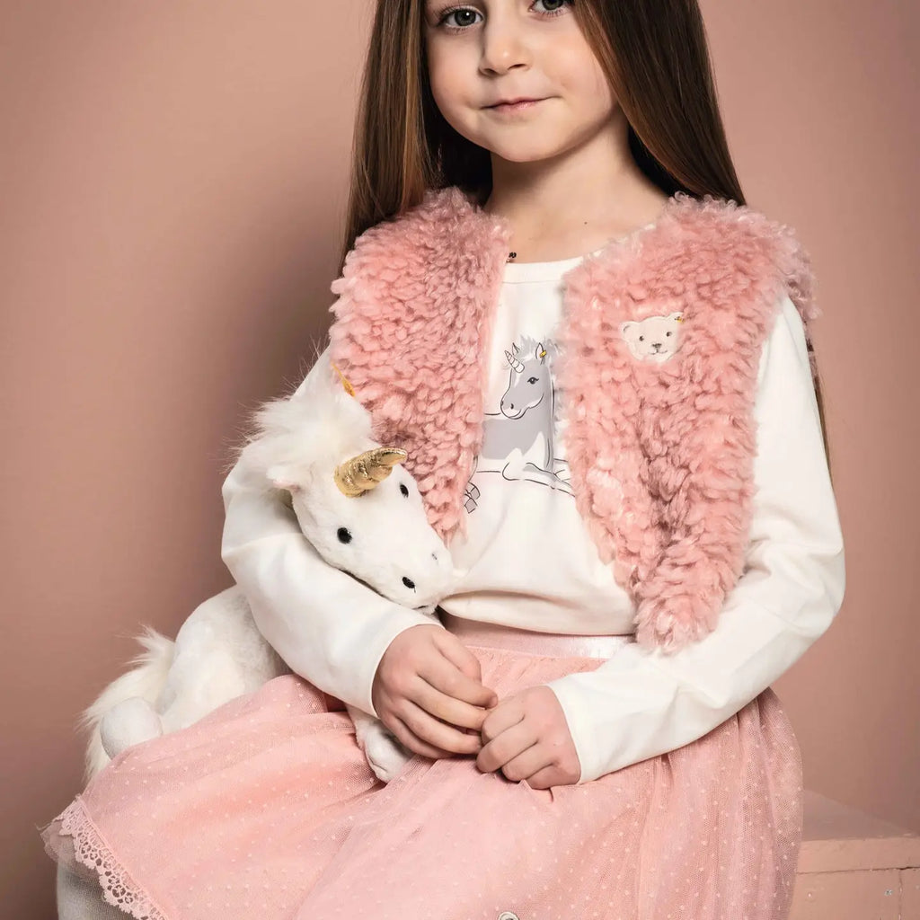 A young girl smiling gently, wearing a pink fluffy vest over a unicorn-themed shirt, holding a Steiff unicorn plush in her lap against a pink backdrop.
