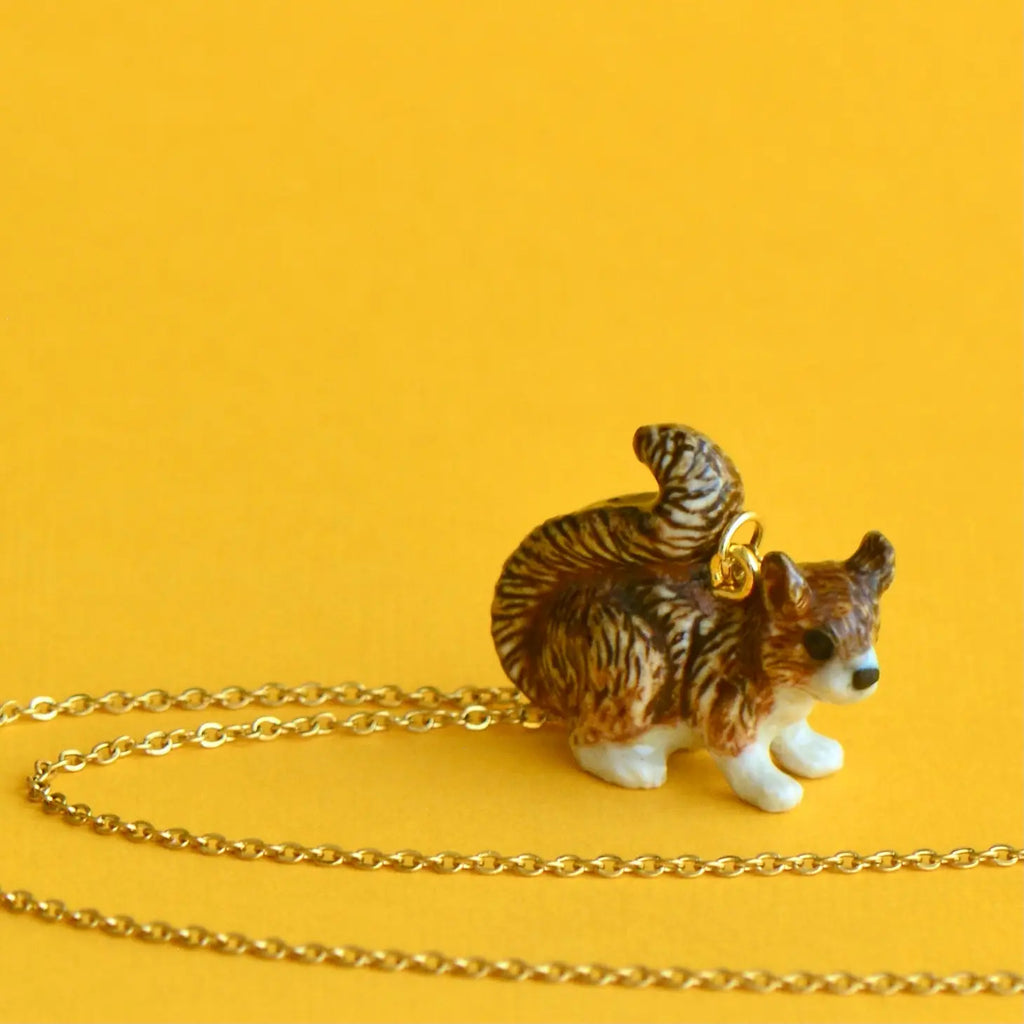 A small porcelain figurine of a brown and white cat, hand painted, with a 24k gold plated Squirrel Necklace attached, sitting on a plain yellow background with a golden chain loosely laid next to
