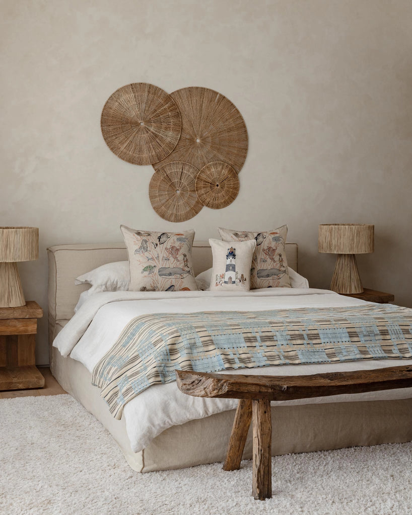 A cozy bedroom with a beige bed, decorative pillows including the Coral & Tusk Lighthouse Friends Pocket Pillow, a blue and beige patterned throw, rustic wooden bench, matching nightstands with woven lamps, circular woven wall art above the bed, and a soft white rug.