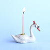 A decorative Swan Cake Topper shaped like a swan with a lit wick, placed against a soft blue background. The swan, a hand-painted porcelain piece, wears a small, red and white striped