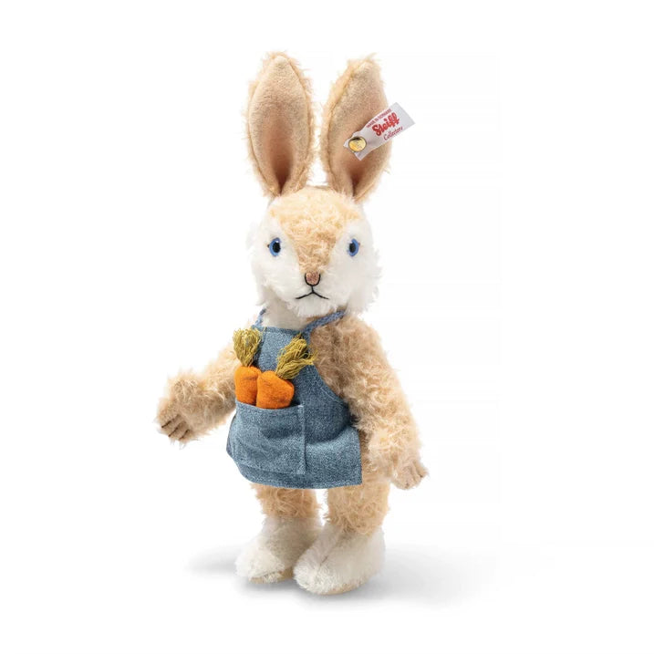 A plush Steiff Carrie Springtime Easter Rabbit standing upright, wearing blue overalls with a small orange carrot stitched onto its pocket, set against a plain white background.