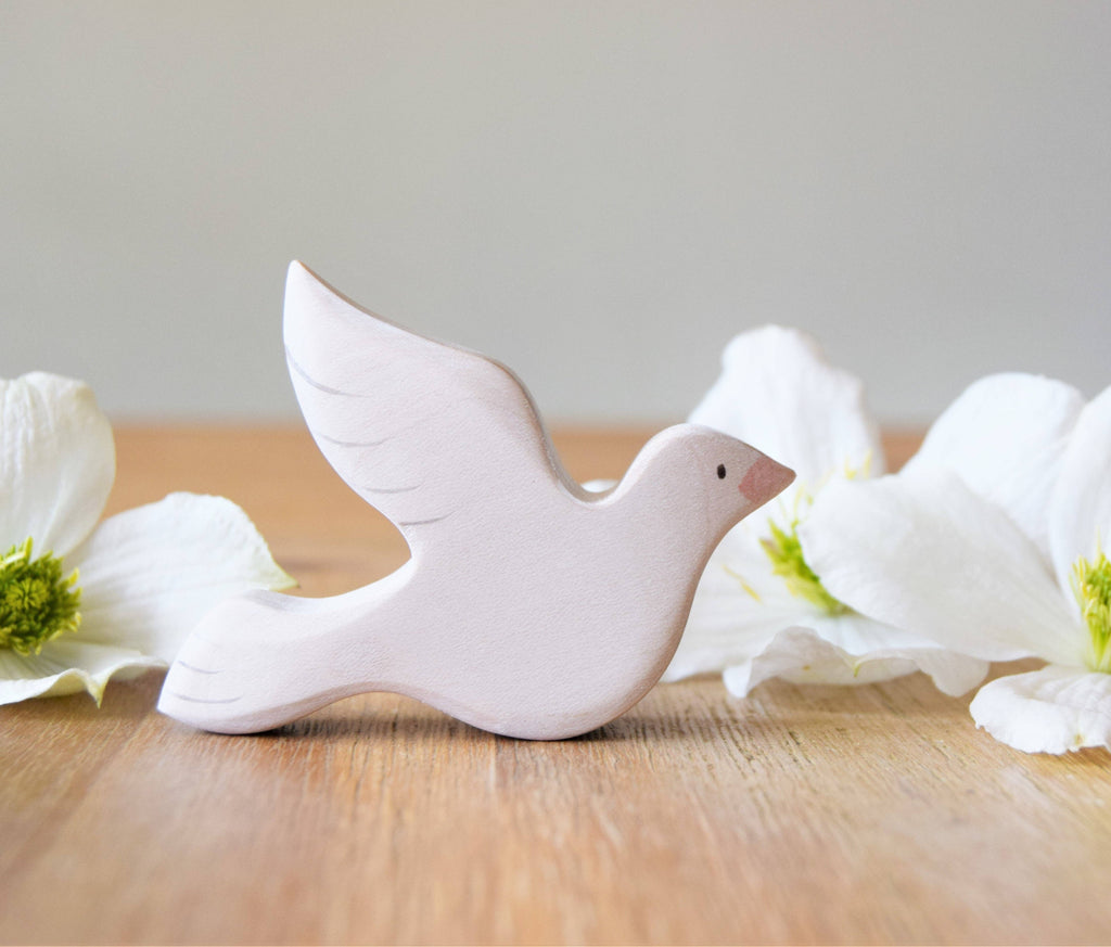The Handmade Wooden Dove, crafted from sustainable hardwood with a minimalist design, sits on a wooden surface surrounded by white flowers. This arrangement creates a serene and peaceful composition, making it perfect for any bird collection.