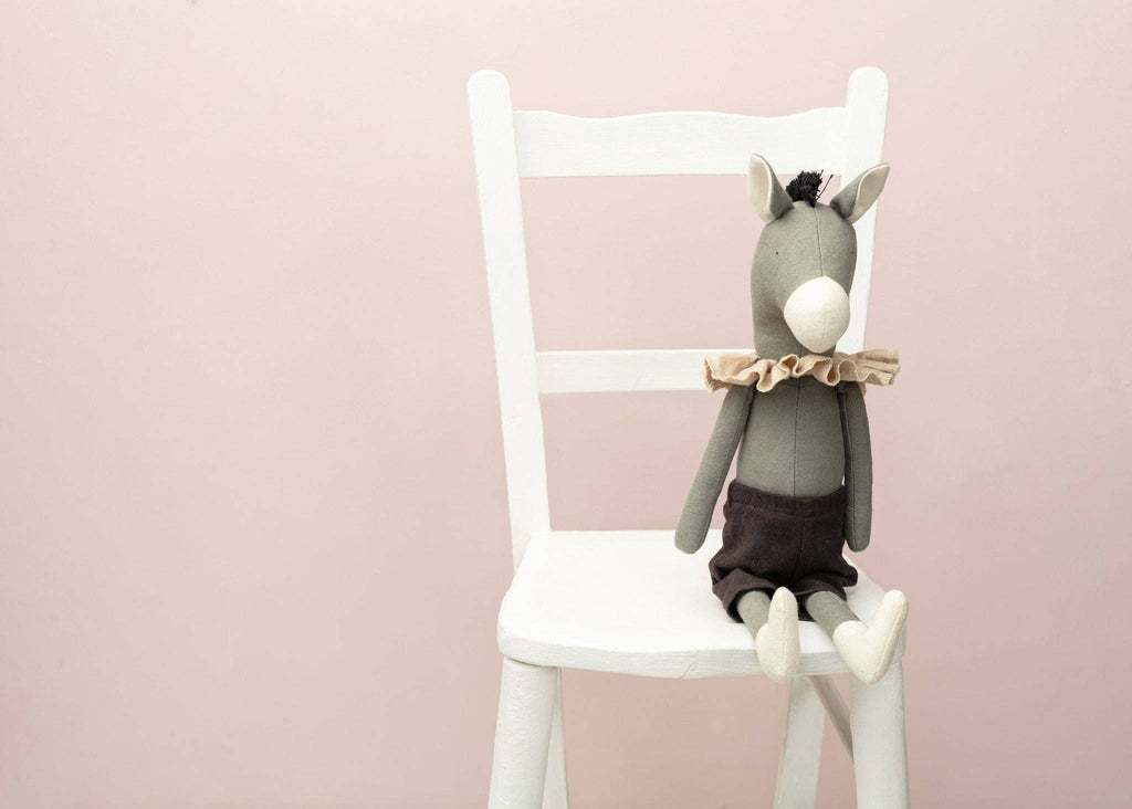 Introducing Darwin the Donkey, a charming gray stuffed toy made from recycled polyester, featuring a ruffled collar and black shorts. It sits on a white wooden chair against a light pink background.