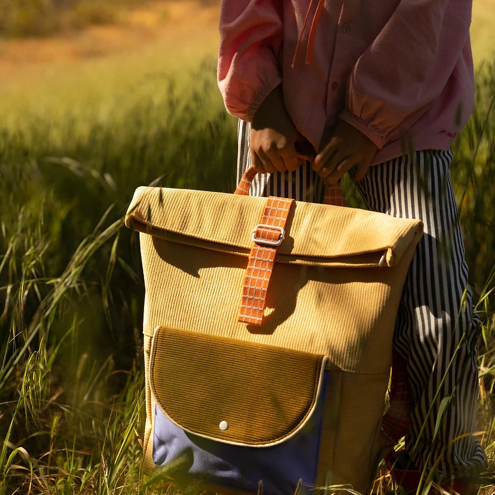A person in striped pants and a pink shirt holds a large, stylish Sticky Lemon Backpack with leather straps in a sunlit field.