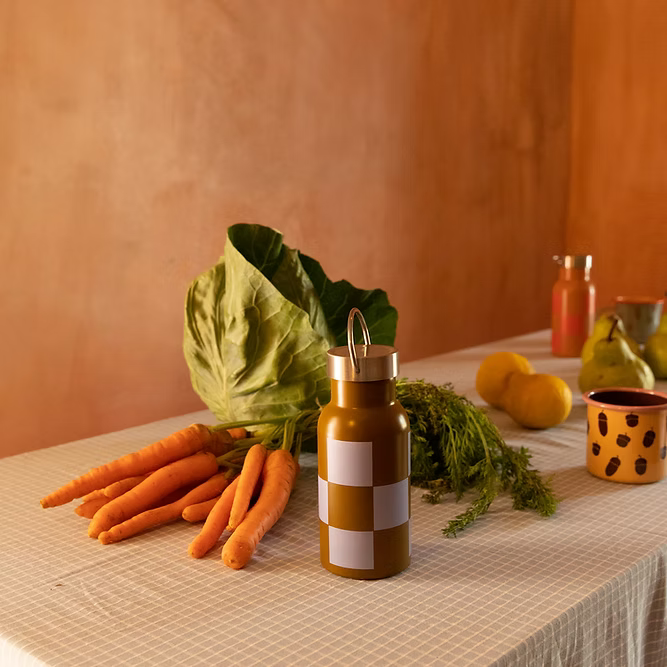A table displays vegetables, fruits, a spotted mug, a glass of juice, and the Sticky Lemon Water Bottle | Farmhouse | Soil Green + Blooming Purple with straw lid against a brown wall.