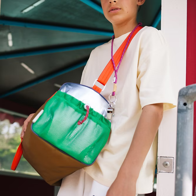 A person in a light-colored shirt holds the Sticky Lemon Sports Bag | Better Together, featuring green, brown, and silver panels with a bright orange strap. The retro bag is made from recycled PET bottles and is held as they look slightly away from the camera.