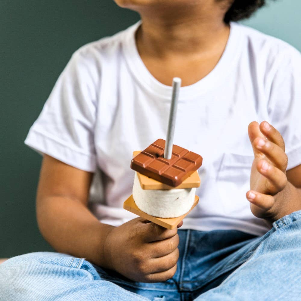 A child in a white t-shirt and jeans holds the Wooden Camp Fire Set s'more on a stick, complete with chocolate, marshmallow, and graham cracker pieces—ideal for indoor camping or kids' pretend play. The child's face is partially out of frame.