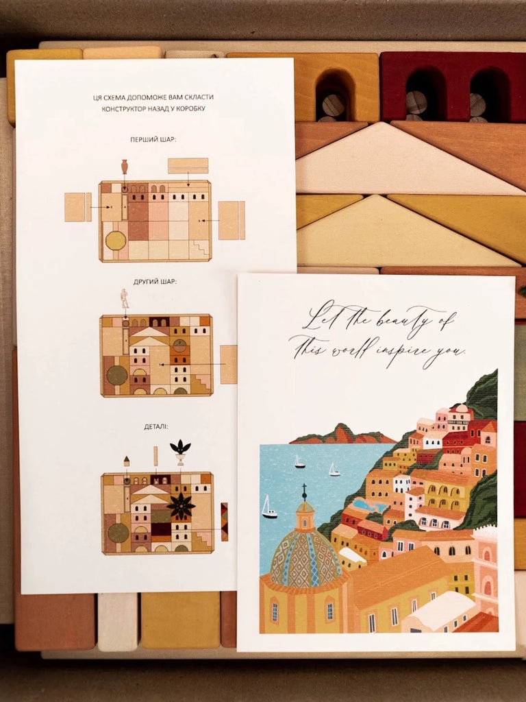 An open notebook displaying Sabo Concept Italian Ancient City Blocks, alongside a card with a picturesque painting of Italian seaside towns. The pages rest on a wooden surface with drawing tools nearby.